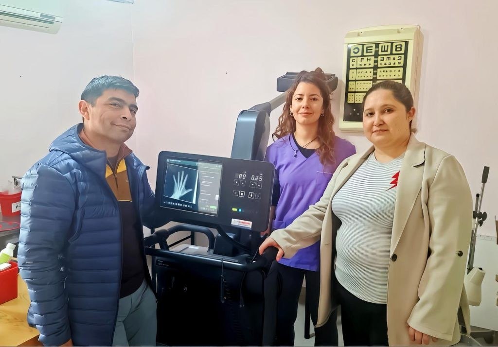 Tres personas se encuentran de pie, una al lado de la otra, frente a una máquina de imágenes médicas. La persona de la izquierda es un hombre con un chaqueta de paja azul sobre una camisa oscura y cabello gris, que mira directamente a la cámara.